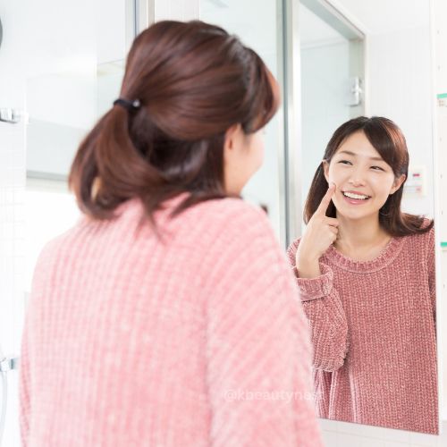 A woman looking into the mirror, smiling, gently patting a clear, watery essence onto her cheeks.